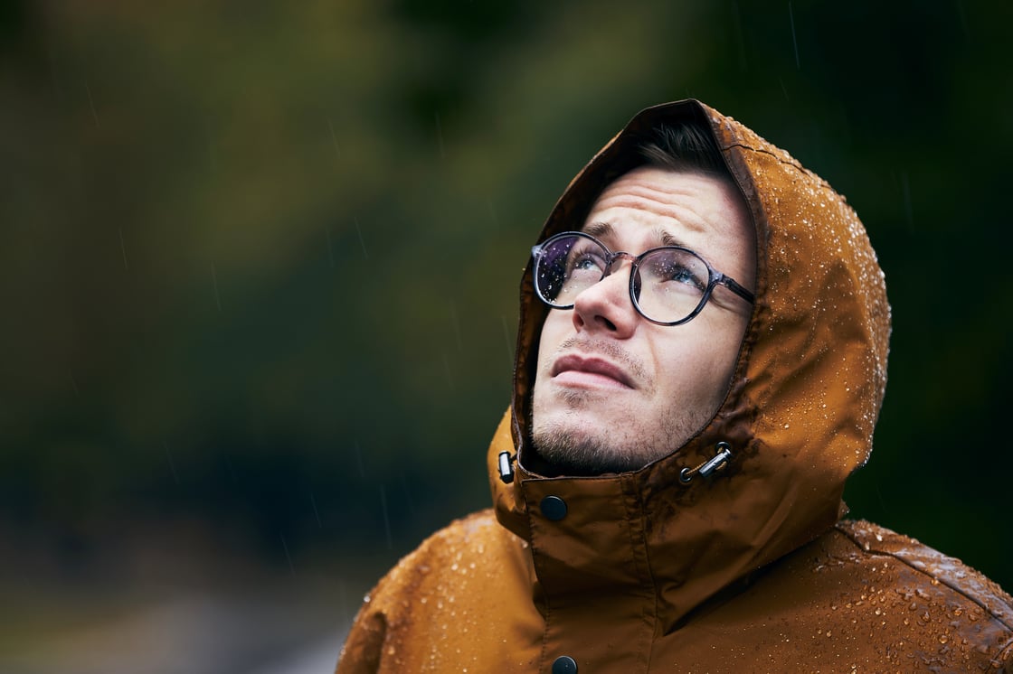man in dripping rain jacket looking up at the sky with a concerned expression